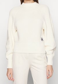 Maglione in maglia crema con collo alto a coste, polsini a coste e orlo aderente. Presenta un logo prominente sul lato.