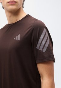 Νεαρό άτομο που φοράει σκούρο καφέ αθλητική κοντομάνικη μπλούζα με τρεις ανοιχτόχρωμες αντανακλαστικές ρίγες στο στήθος και το μανίκι.