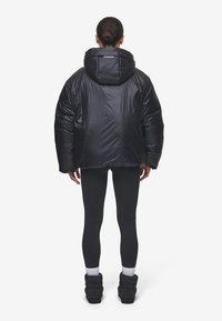 Zwarte gewatteerde jas met capuchon, voorzien van een glanzende afwerking en een oversized ontwerp. Draagt met strakke zwarte leggings en enkellaarsjes.