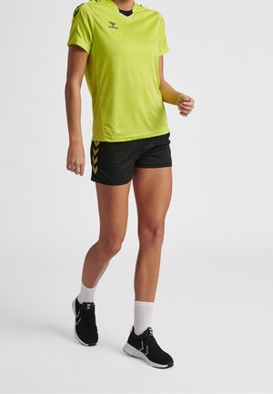 Persona que lleva una camiseta deportiva verde brillante, pantalones cortos negros, calcetines blancos y zapatos deportivos negros sobre un fondo claro liso.