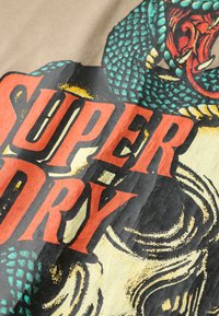 Koszulka graficzna z wyrazistym czerwonym napisem "SUPER DRY" na kremowym tle, z zielonym i czerwonym wzorem węża oraz teksturowanym materiałem.