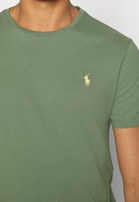 Oliivinvihreä puuvillainen T-paita, jossa on pyöreä pääntie, lyhyet hihat ja pieni kultainen kirjailtu polohevosen logo rinnassa.