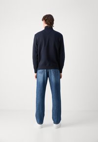 Navy sweatshirt with a high collar, paired with light blue wide-leg jeans and white sneakers, viewed from the back on a neutral background.