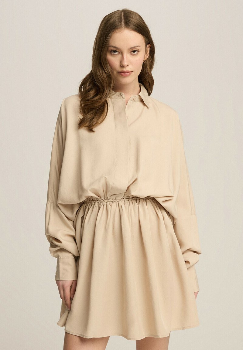 Beżowa sukienka koszulowa z zapięciem na guziki przy kołnierzy, długimi rękawami i elastycznym pasem, charakteryzująca się luźnym krojem i gładką fakturą materiału.