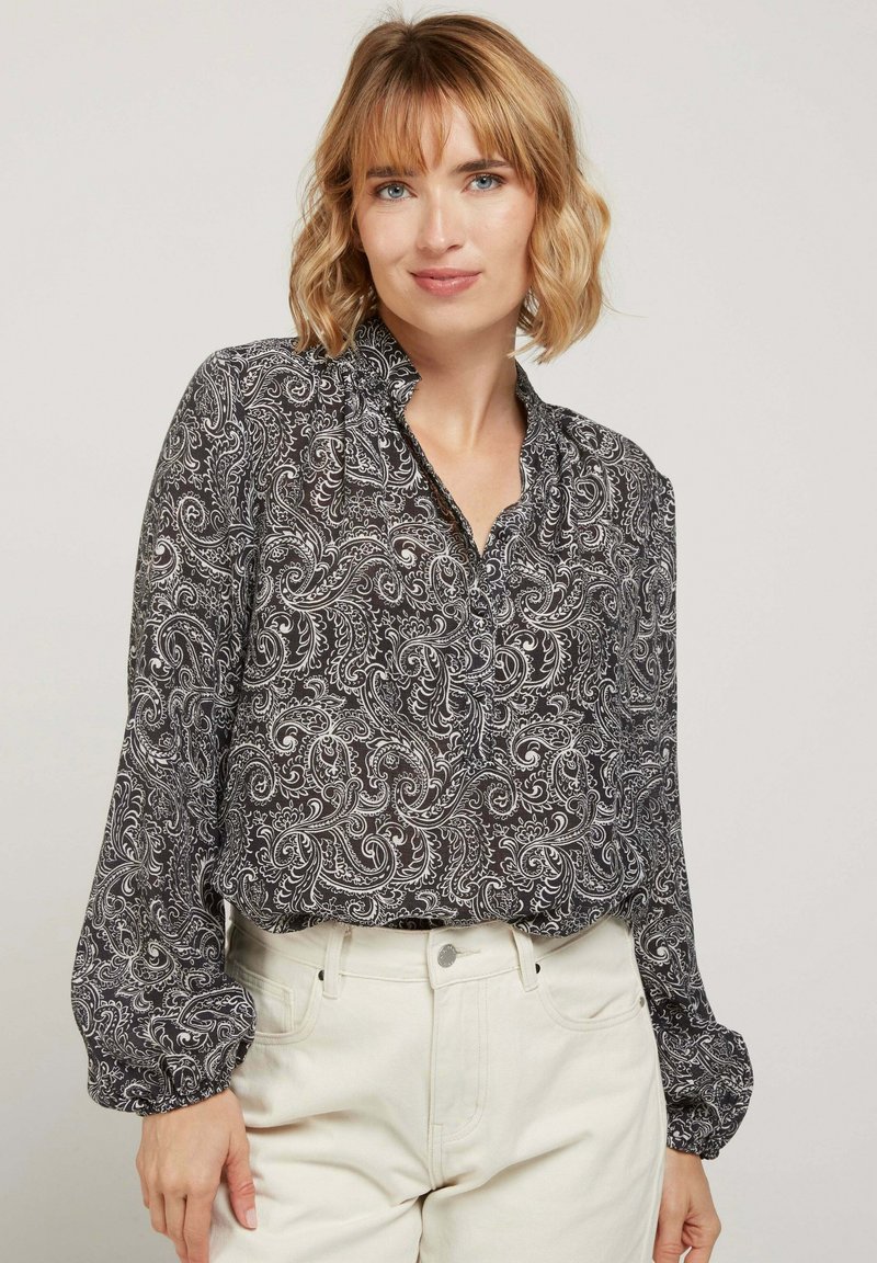 Blouse paisley noir et blanc avec un col volanté, des manches longues bouffantes et une coupe décontractée, accompagnée d'un pantalon de couleur claire.