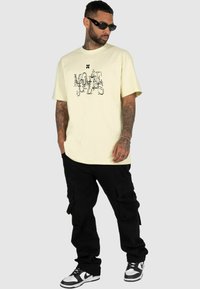 Yellow cotton t-shirt with graphic print, paired with black cargo pants featuring pockets. Footwear is black and white sneakers.