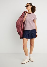 Γυναίκα που φοράει γυαλιά ηλίου, ριγέ μπλουζάκι, τζιν φούστα, λευκές κάλτσες με αθλητικά παπούτσια, κρατώντας ένα κόκκινο μπουφάν στον ώμο της μπροστά σε απλό φόντο.