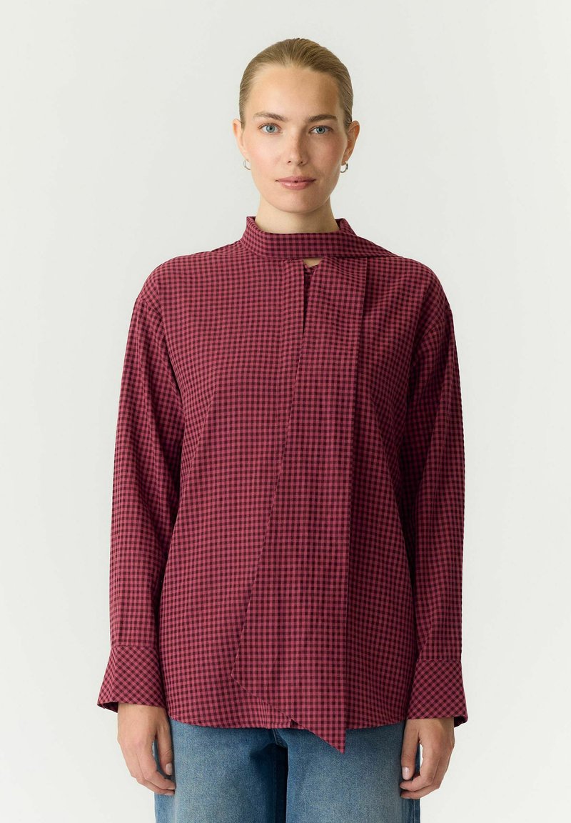 Femme portant un chemisier à manches longues à carreaux rouges et noirs avec un détail cravate, assorti à un jean bleu, debout devant un fond uni.