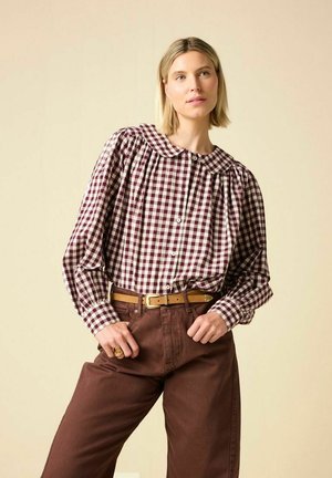 Vrouw draagt een bordeauxrode en witte geruite blouse met een ronde kraag, bruine hoge taille broek en een beige riem, poserend tegen een beige achtergrond.