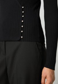 Haut à manches longues noir côtelé avec un col en V et de boutons décoratifs en argent sur le côté. Associé à un pantalon noir en tissu lisse.
