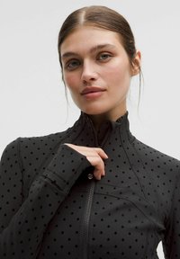 Chaqueta negra de cremallera con una textura suave y un patrón de lunares en relieve. Cuenta con un alto cuello y mangas alargadas con orificios para los pulgares.