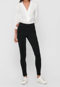 Jeans negros de talle alto con corte ajustado, hechos de denim. Combinados con una camisa blanca de manga larga y zapatillas blancas.