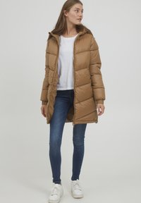 Manteau en duvet marron avec capuche, présentant un design matelassé, une fermeture éclair et des poignets côtelés. Associé à un jean foncé et des baskets blanches.