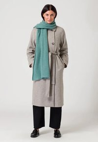 Abrigo de lana gris con cintura ceñida y cierre de un solo botón. Combinado con una bufanda de textura en color teal y pantalones negros de pierna ancha.