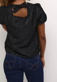 Blouse noire texturée à manches courtes avec manches bouffantes et détail nœud au dos, portée avec un jean bleu foncé.