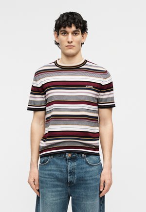 Άνδρας φορώντας πολύχρωμο κοντομάνικο πουκάμισο με οριζόντιες ρίγες και μπλε τζιν, στέκεται μπροστά σε απλό φόντο.