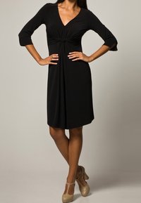 Schwarzes Kleid mit ¾-Ärmeln, V-Ausschnitt und geknotetem Detail in der Taille. Gerader Rock, glatte Textur, kombiniert mit hellen, hohen Schuhen.