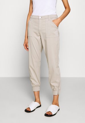 Beige cargobroek met elastische boorden, twee voorzakken en een knoopsluiting, gecombineerd met witte slippers met kruisbanden. Glad textuur.