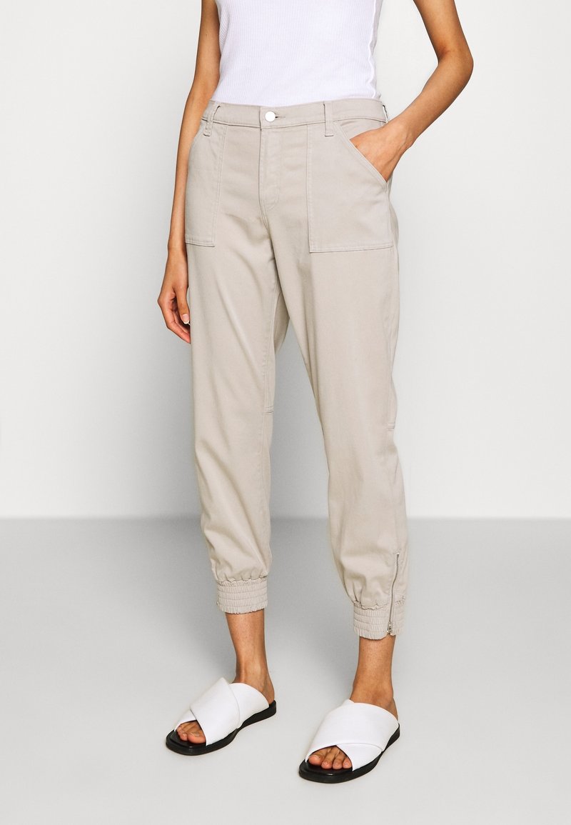 Beige cargo kalhoty s elastickými manžetami, dvěma předními kapsami a zapínáním na knoflík, spárované s bílými sandály se zkříženými pásky. Hladká textura.