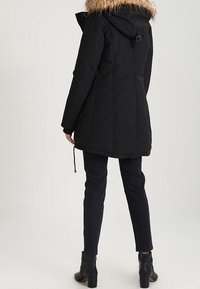 Zwarte geïsoleerde parka met een met bont gevoerde capuchon, taille met trekkoord en zijzakken, gecombineerd met donkere broek en enkelboots.