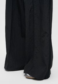 Μαύρο παντελόνι φαρδιάς γραμμής που καλύπτει μερικώς ασημένια παπούτσια με ανοιχτό καφέ σόλες σε ανοιχτό φόντο.