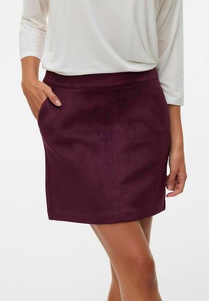 Vrouw met een donkerpaarse suède minirok met een elastische tailleband en een wit, langärmelig, ingestopt shirt, met één hand in de zak van de rok.