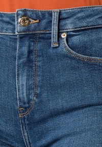 Jeans de denim azul oscuro con herrajes de bronce, que cuentan con cierre de botón, costuras en contraste y bolsillos frontales. Vista en primer plano de la tela.