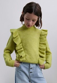 Suéter verde lima con acentos con volantes en los hombros, cuello y puños acanalados, combinado con jeans de tiro alto en azul claro.