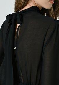 Schwarze Bluse aus durchscheinendem Stoff, mit hohem Kragen und einer Schleifenverschluss hinten. Bemerkenswertes Knopfdetail unterhalb des Ausschnitts.