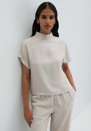 Frau mit langen, dunklen Haaren, die ein beiges, kurzärmeliges Oberteil mit hohem Kragen und passende Hosen trägt, steht vor einer hellen Wand.