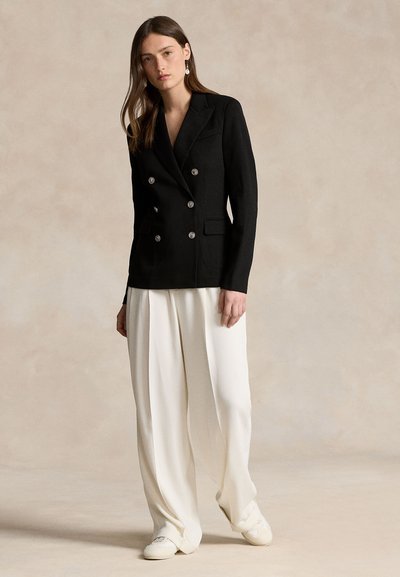 Femeie în picioare, îmbrăcată cu un sacou negru cu două rânduri de nasturi, pantaloni albi cu picioare largi, adidași albi și o cercei lungi, pe un fundal neutru.