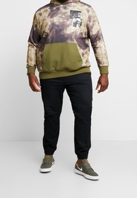 Mikina s kapucí s tie-dye potiskem v hnědé a olivové zelené, se přední kapsou a bílým logem. Spárováno s černými tepláky.