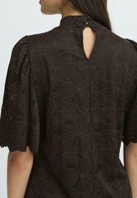 Brun bluse med blomsterlaceringsdesign, høj halsudskæring, korte puff�ærmer og en bageste nøglehulåbning med en diskret knaplukning.