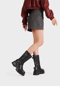 Stivali da combattimento in pelle nera con suola spessa, caratterizzati da fibbie argentate e una finitura testurizzata. Indossati con una mini gonna grigia.