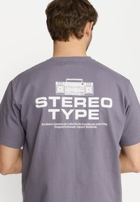 Grijze katoenen t-shirt met een witte grafische afbeelding van een boombox en de tekst "STEREO TYPE" op de achterkant. Korte mouwen, ronde halslijn.