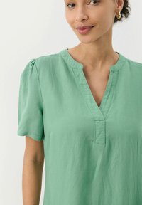 Kurzarm, V-Ausschnitt-Oberteil aus weichem grünen Baumwollstoff. Mit gefältelten Schultern und einem entspannten Schnitt. Sauberes Design mit minimalen Details.