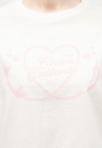 Λευκό μπλουζάκι με σχέδιο ροζ τσίλι σε σχήμα καρδιάς και κείμενο που γράφει "Chica Caliente" στο στήθος.