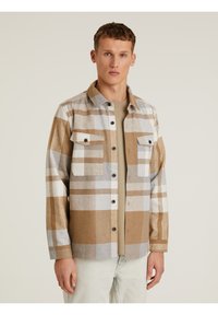 Camisa de franela a cuadros marrón y gris con dos bolsillos frontales, cierre de botones y cuello; combinada con una camiseta beige clara debajo.