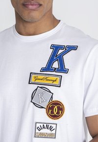 Bijela pamučna majica s više izvezenih zakrpa: plavi "K", žuti "Gianni Kavanagh", sivi "K" i crveni "GG" logotipi.