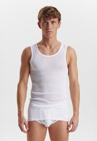 Hvid ribbet tanktop lavet af bomuld, med rund hals og brede stropper. Båret over hvide trusser, med en tætsiddende pasform.