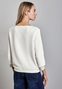 Weißer strukturierter Pullover mit weitem Ausschnitt und Dreiviertelärmeln, kombiniert mit blauen Jeans. Der Pullover hat einen gerippten Saum.