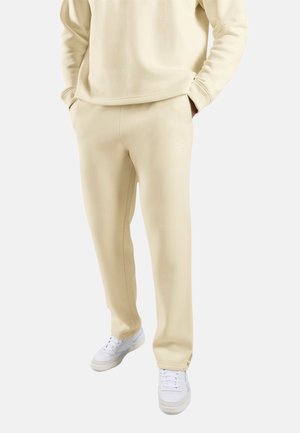 Persoon draagt beige sweatpants en bijpassende sweatshirt met handen in de zakken, gecombineerd met witte sneakers op een effen achtergrond.