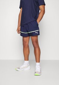 Navy urheilushortsit neonvihreillä yksityiskohdilla ja Puma-logolla, yhdistettynä valkoisiin ja monivärisiin tennareihin, joissa on vihreä pohja.