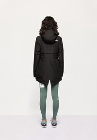 Giacca impermeabile nera con cappuccio, caratterizzata da una vita cinte e un retro più lungo. Indossata sopra leggings verdi e scarpe da corsa.