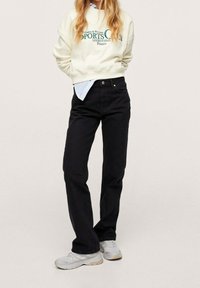 Persona che indossa un felpa crema con testo verde, jeans neri a vita alta, una maglietta a righe che sbircia, e scarpe da ginnastica beige, in piedi su uno sfondo chiaro.