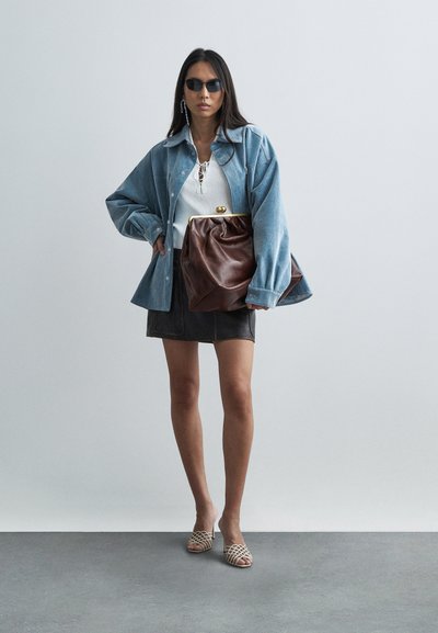 Mujer con gafas de sol negras, chaqueta azul clara de gran tamaño, top blanco, minifalda negra, tacones beige abiertos en los dedos, sosteniendo un gran bolso de mano de cuero marrón.