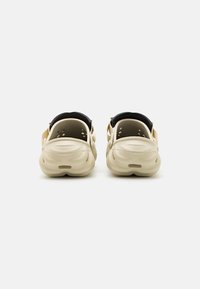 Sabots beige à enfiler avec des trous de ventilation, semelles texturées et brides de talon rembourrées noires, vus de dos sur un fond uni.