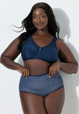 Pewna siebie kobieta z długimi włosami pozująca w granatowym biustonoszu z koronkowymi akcentami i dopasowanych majtkach z wysokim stanem i wzorem.