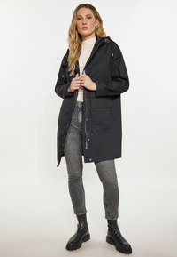 Parka nera impermeabile con cappuccio, chiusura a zip frontale e bottoni a scatto. Presenta ampie tasche frontali e una vestibilità rilassata; abbinata a jeans grigi e stivali.