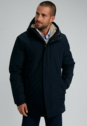 Homme portant un manteau d'hiver à capuche bleu marine sur une chemise bleu clair, se tenant les mains dans les poches, regardant sur le côté devant un fond uni.